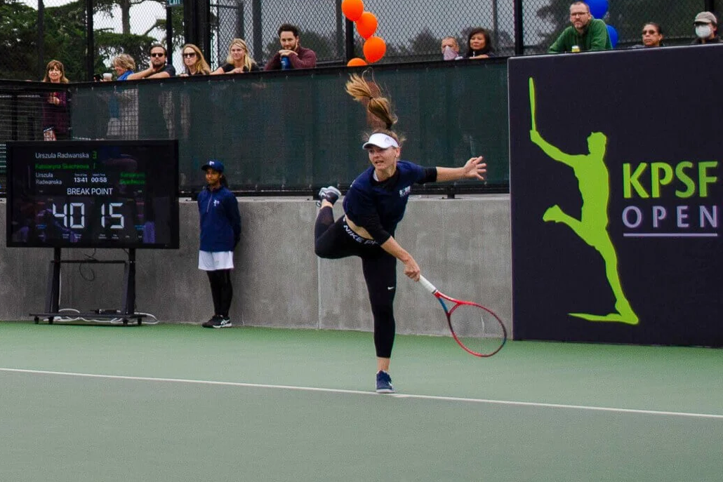 Player serving on centre court at the KPSF Open with scoreboard and branded backdrop